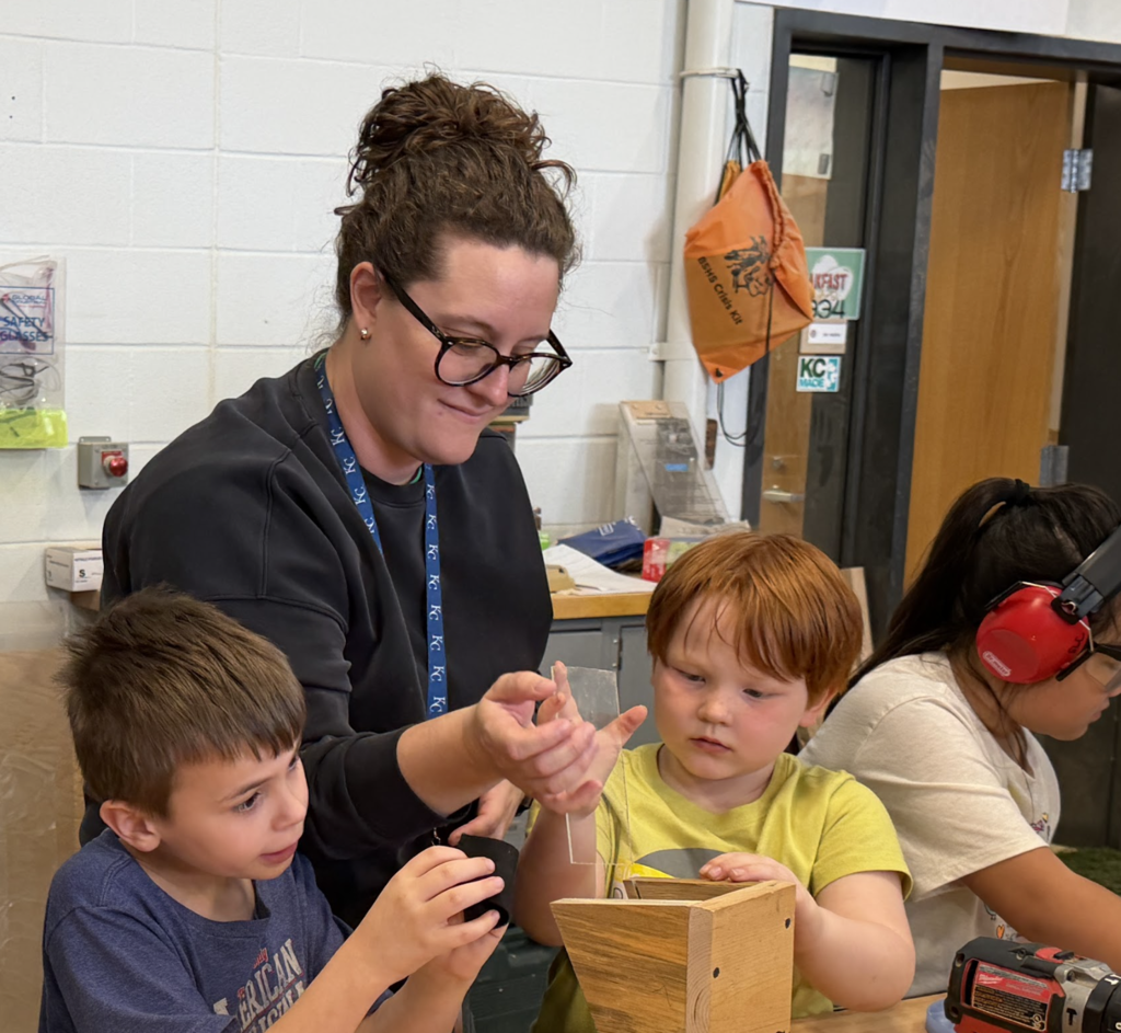 Two boys look at a wooden box while a woman shows them something. Behind them, a woman uses headphones and a drill.