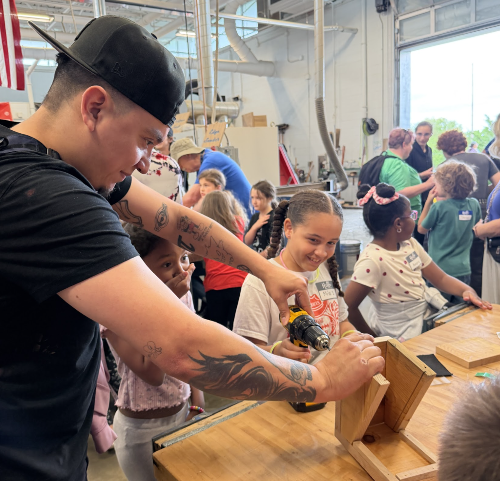 A man helps a girl with a drill while crafting at a table, in a workshop.