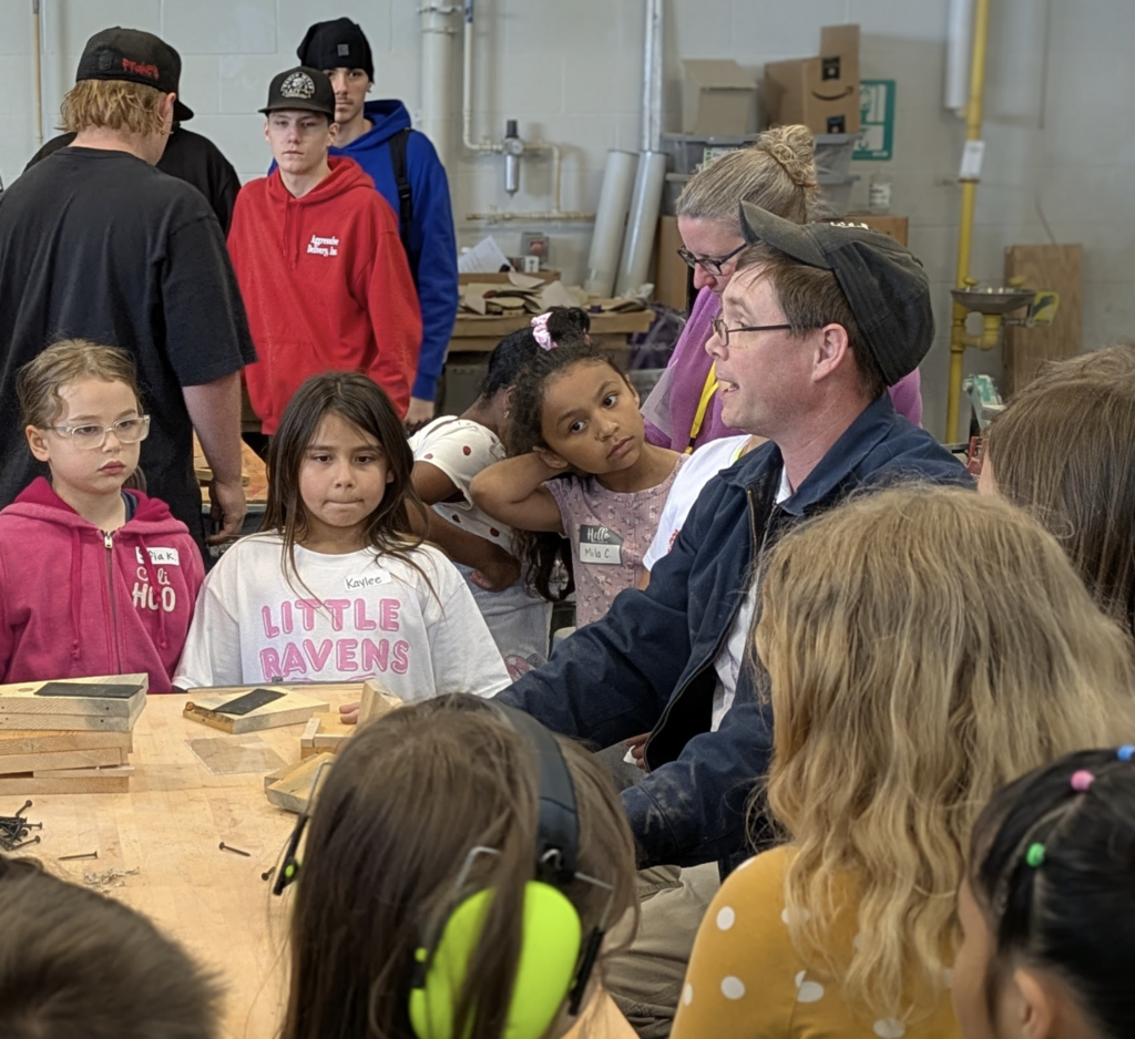 A group of children and adults in a room. A man in a blue jacket stands next to a desk.