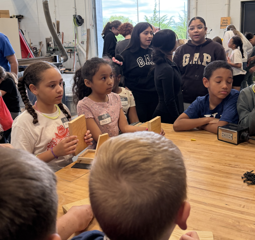 A group of children work on projects at a table, with others observing. The table has wood pieces.