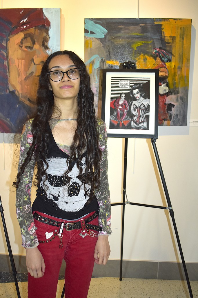 Woman with long dark hair wearing glasses and red pants stands in front of an easel.