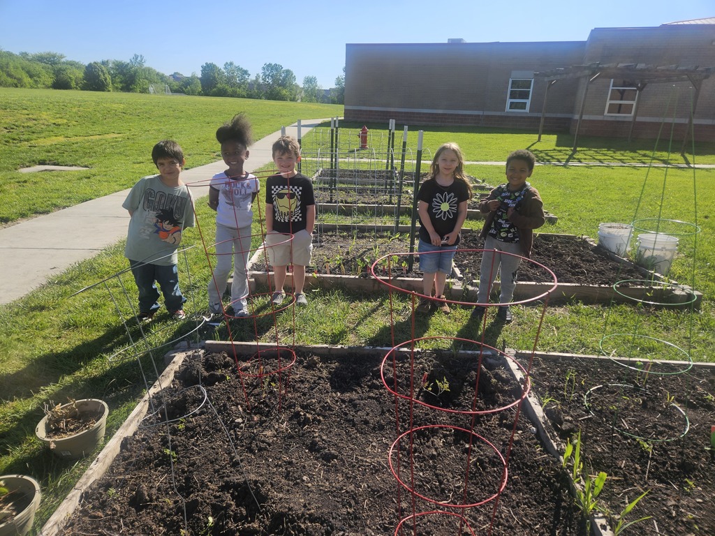 Five children pose in a garden area, with raised beds, red hoops, and a building in the background.