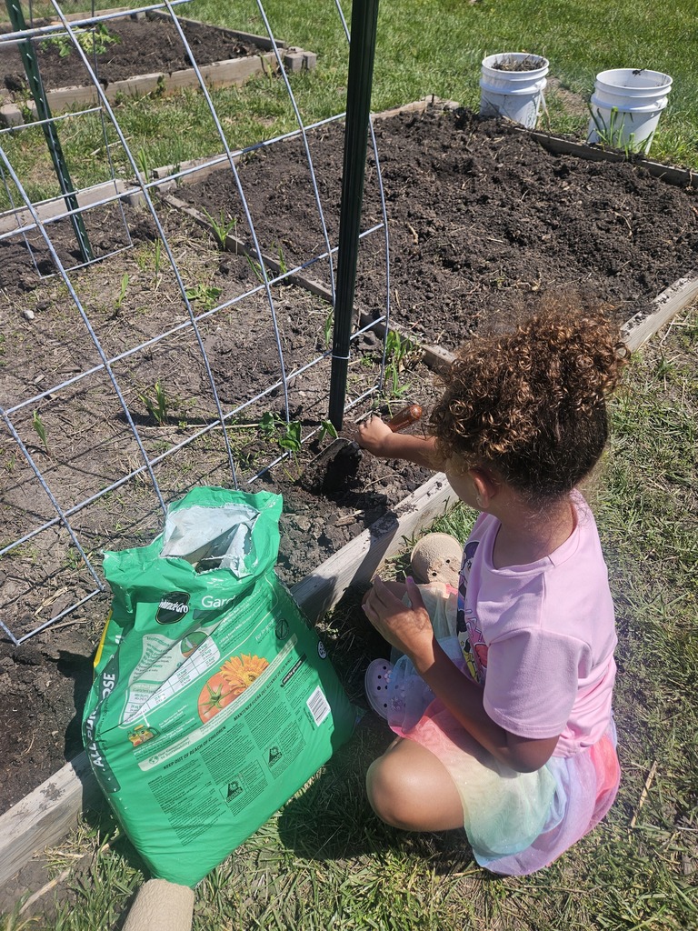 A child sits on grass near a metal support, planting seedlings. A green bag of soil is nearby.