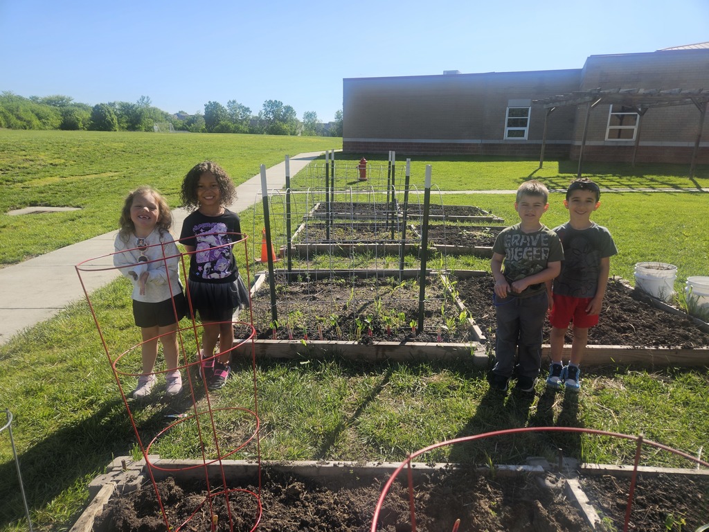 Four children stand in a garden with plants, raised beds, and a building in the background.