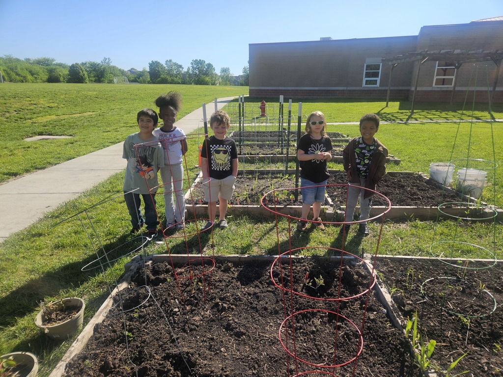 Five children in a garden with raised beds, a school building behind them, and a sunny day.