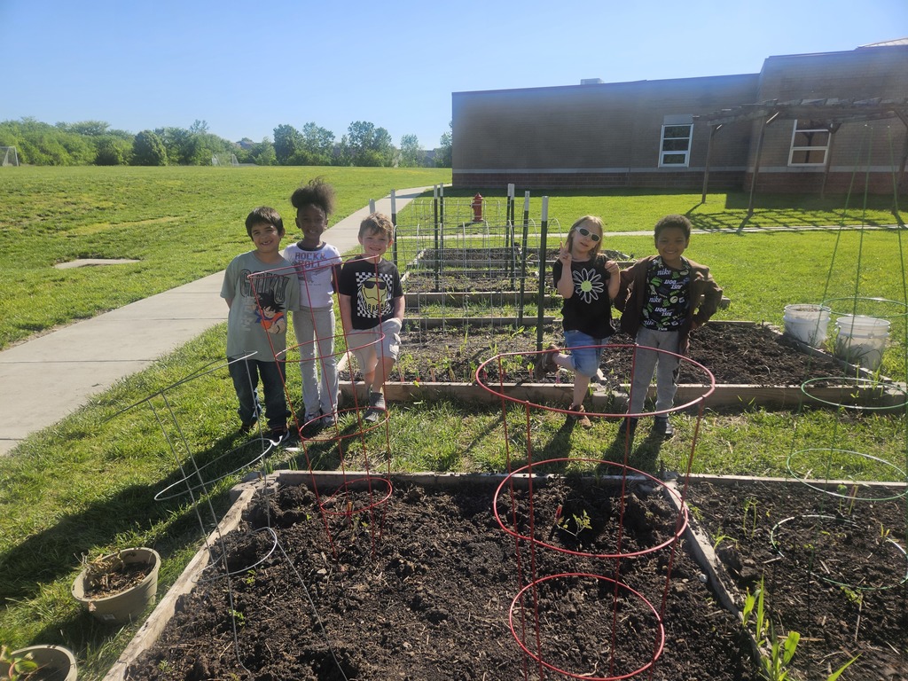 A group of children in a school garden, with raised beds, tools, and a building in the background.