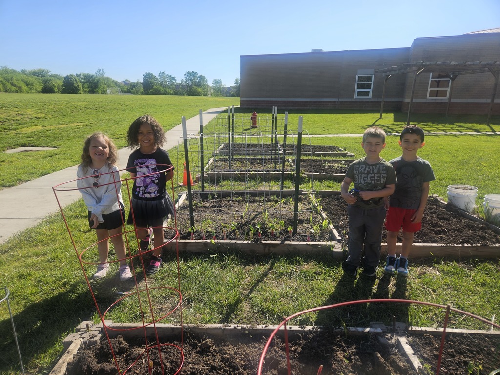 Four children stand in a vegetable garden with raised beds. Behind them, a building and trees are visible.