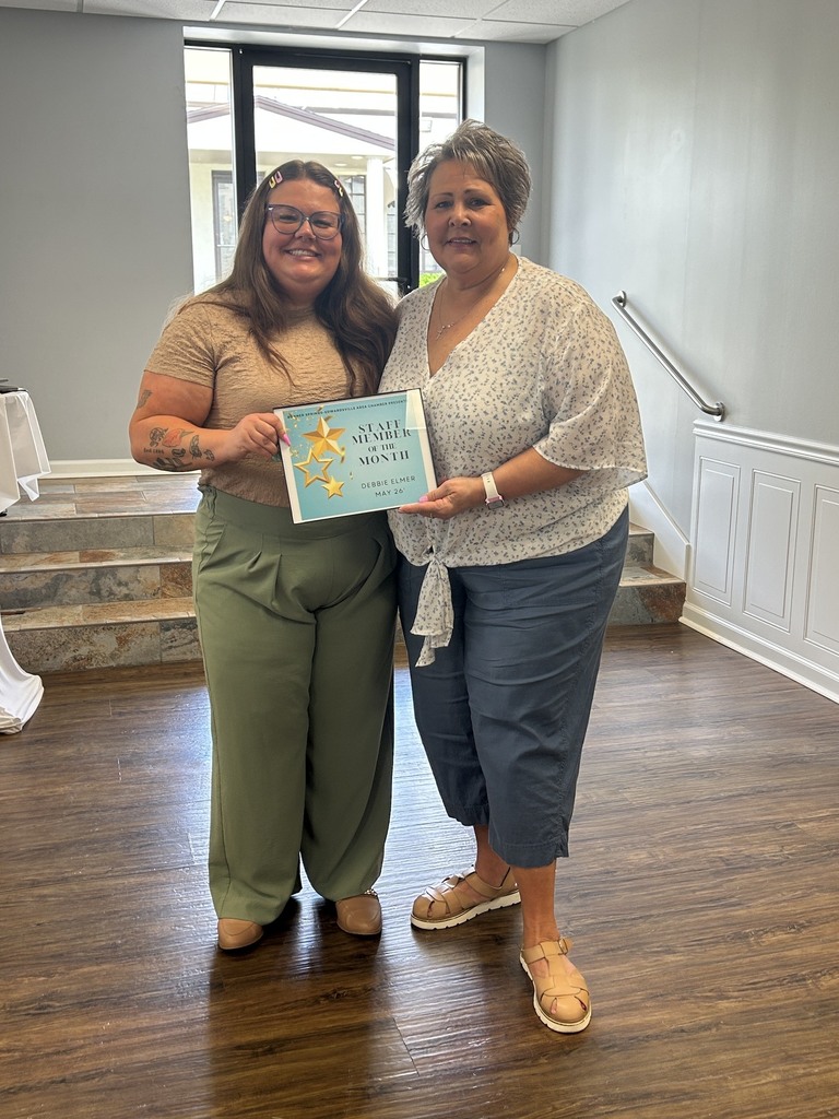 Two women stand on a wooden floor, one holding a certificate. They are in a room with stairs and a glass door.