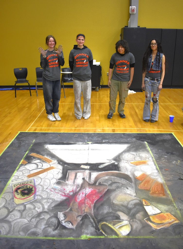 Four individuals stand around a large, multicolored chalk drawing on a floor. The backdrop is a yellow wall.