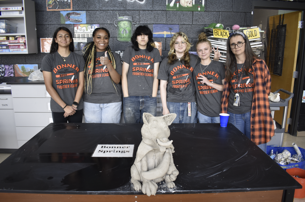 Six people in matching gray t-shirts stand in a room, with one holding a cup. A table has a clay sculpture.