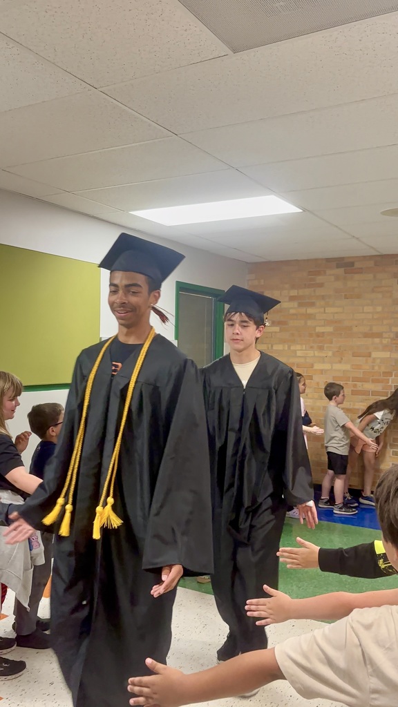 Two men in graduation gowns and caps walk between others. A person in a tan shirt reaches out to touch them.