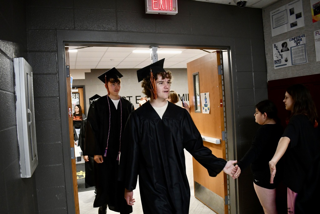 Three students in caps and gowns walk through an open door into a room; another student stands in the background.