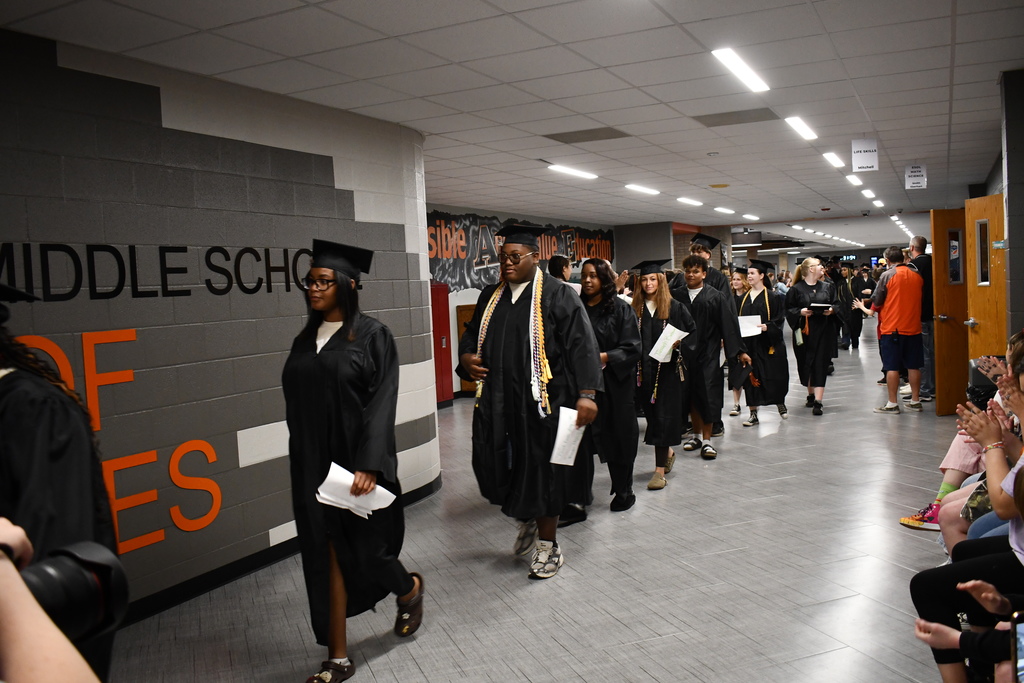 Graduates in caps and gowns walking through hallway. Audience seated on one side.