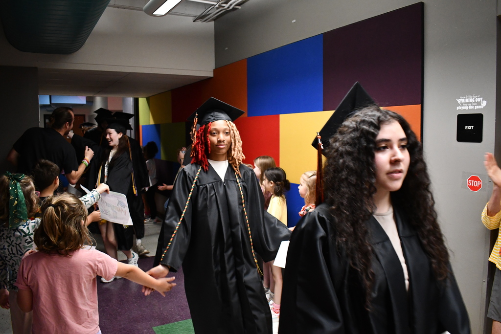 A group of people, including two in graduation gowns, walk through a colorful hallway with children gathered.