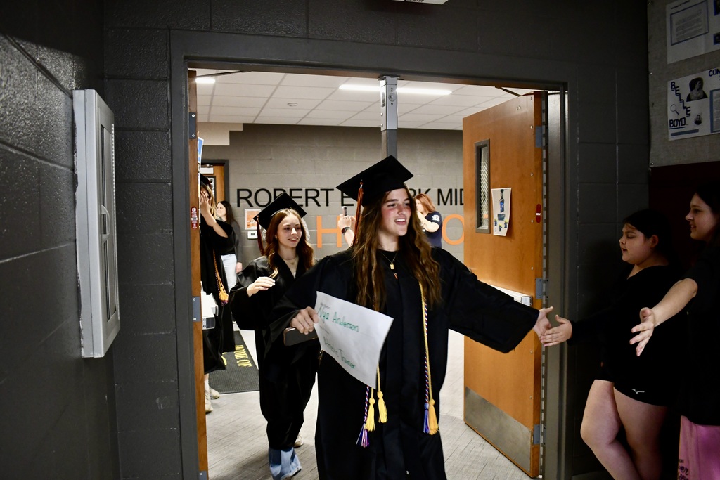 A woman in a graduation gown stands with a paper and cap, facing others in similar gowns near an open door.