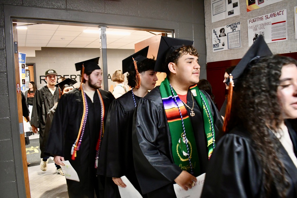 Graduates wearing gowns and caps are lined up in a hallway, some with sashes and others holding papers.
