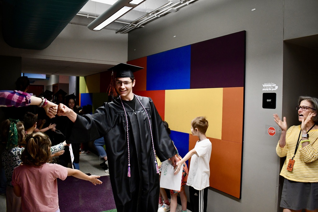 A man in a graduation gown and cap walks with others. Colorful squares decorate the wall behind him.