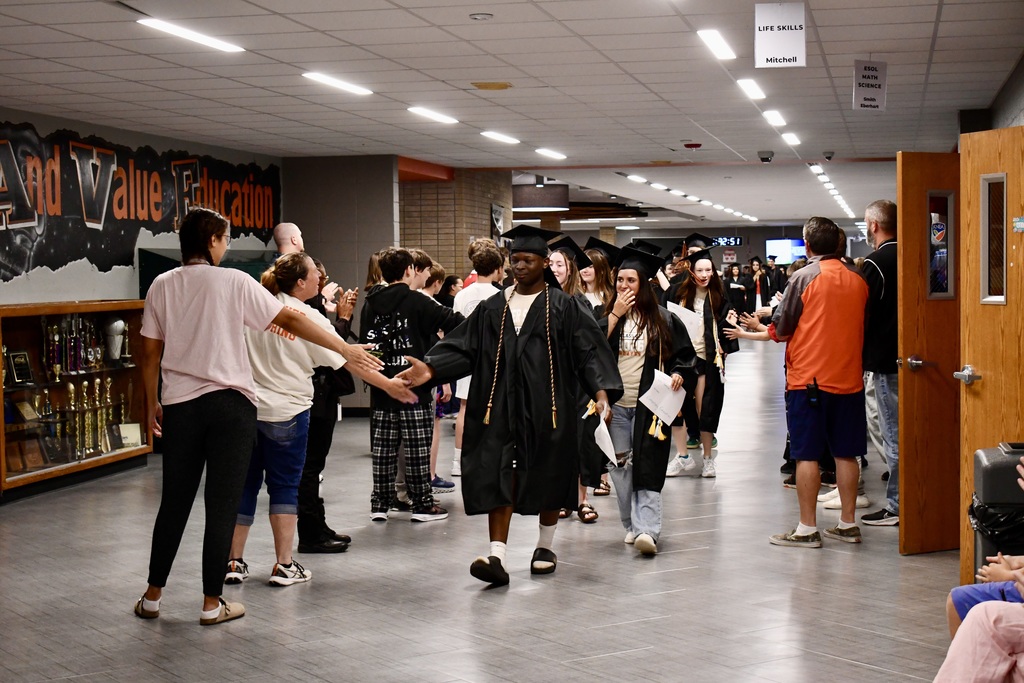 A group of people walk in a hallway, a man in a graduation gown leads, others follow, and a cabinet with bottles is in the background.
