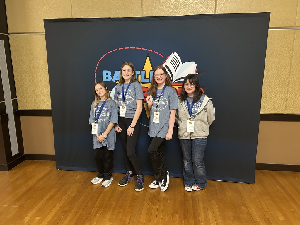 Four girls stand in front of a backdrop with a logo and a book. They wear casual clothes and ID lanyards.