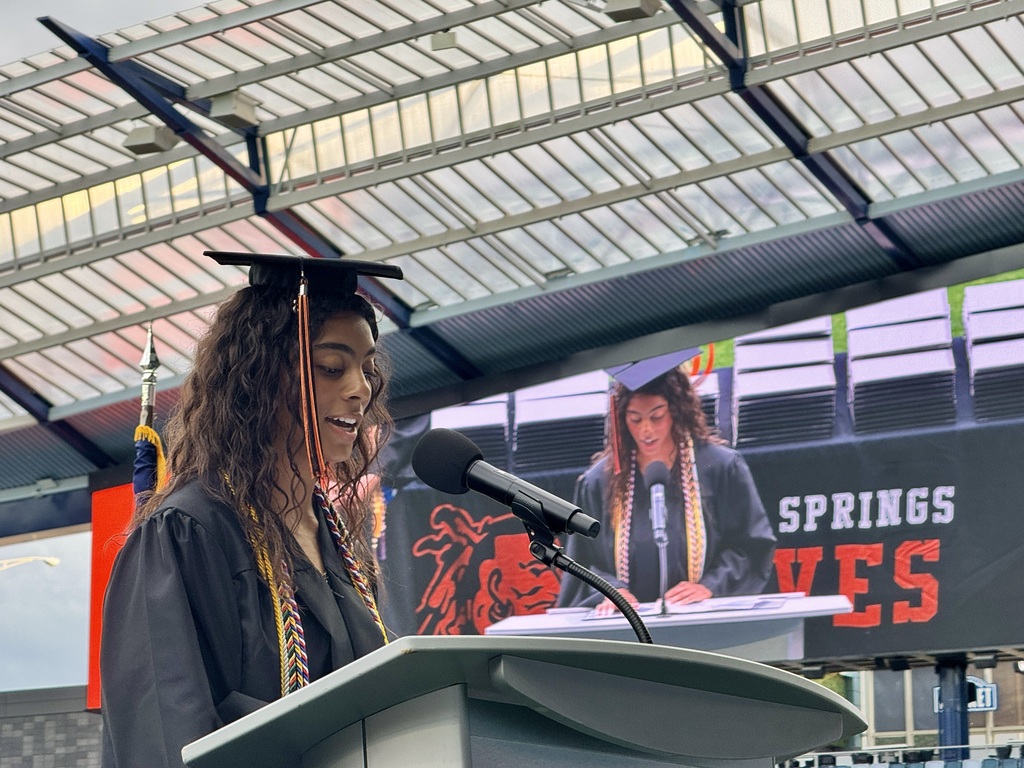 A woman in a cap and gown stands at a podium with a microphone. A large screen behind her displays text.