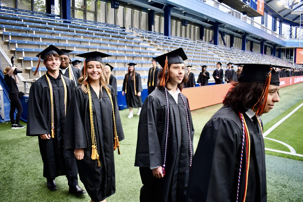 A group of people in black gowns and caps walking on a green field, possibly at a graduation.