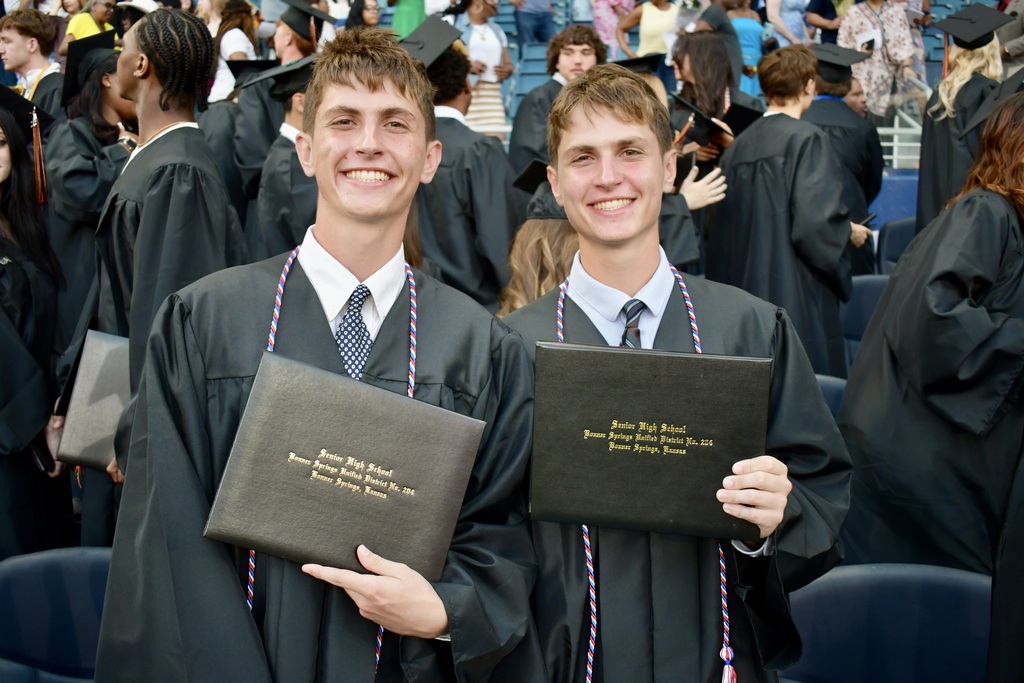 Two men in black graduation gowns with tassels hold up their diplomas at a ceremony.
