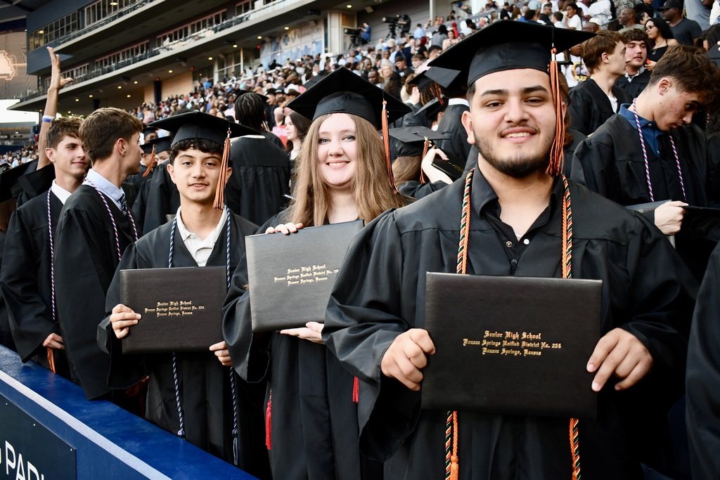 Graduates in academic regalia stand in a stadium, holding diplomas, with spectators in the background.