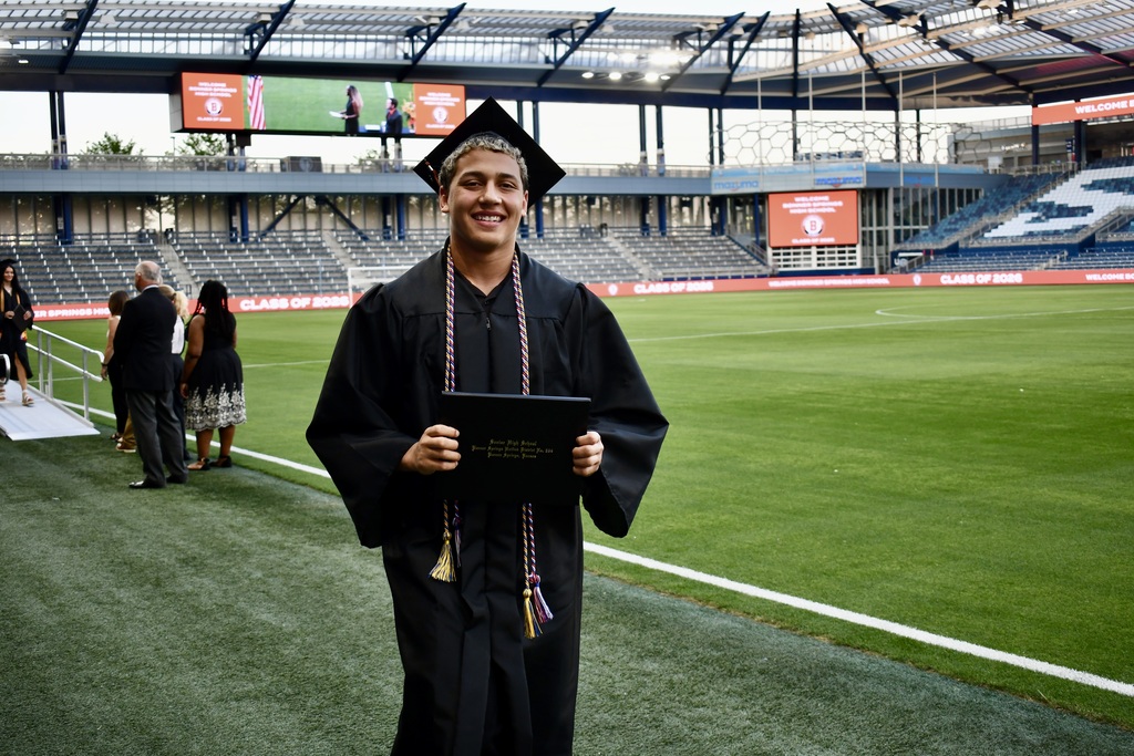 A person in a cap and gown stands in a stadium, holding a diploma, with empty bleachers and a field.
