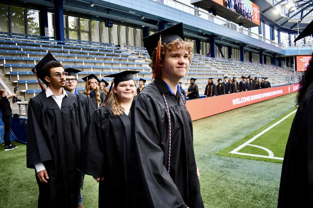 A group of people wearing graduation gowns and hats stand on a field, with bleachers in the background.