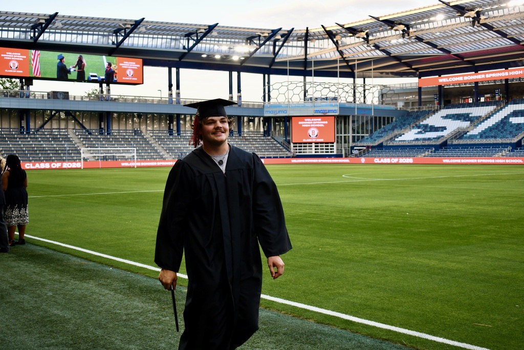 A person in a graduation gown walks across a green field with stadium seating in the background.