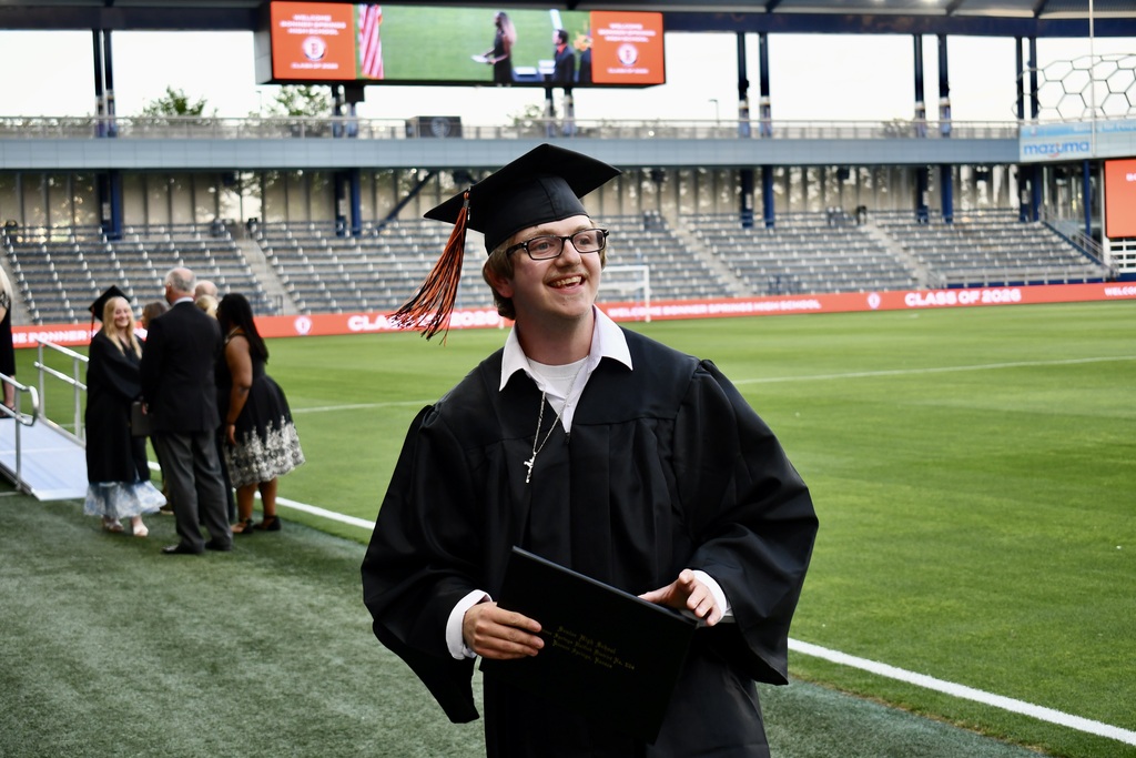 A person in a graduation gown stands on a field with an empty stadium in the background.