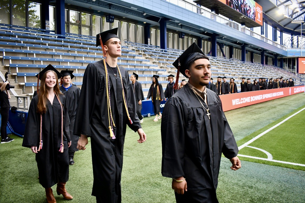 Graduates wearing caps and gowns walk on a field, with empty stadium seats in the background.