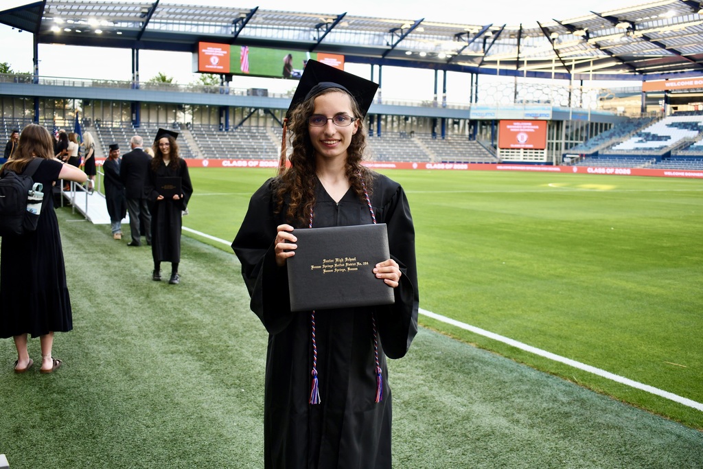 A person in a graduation gown poses with a diploma in front of an empty sports stadium.