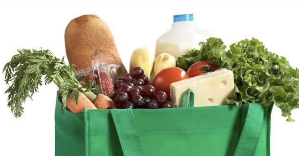 A green bag contains bread, grapes, tomatoes, cheese, carrots, lettuce, milk, and a plastic wrap.