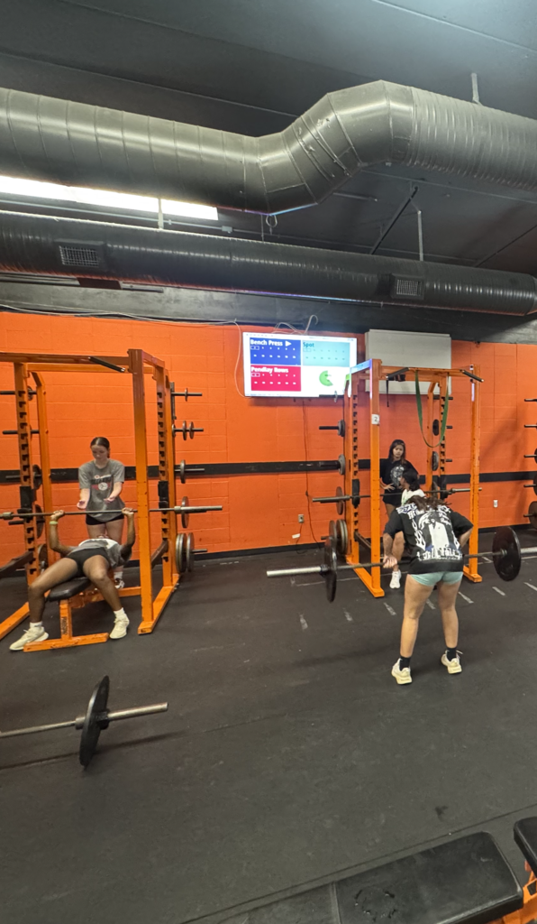 Gym with orange walls. Four people work out. One lifts a barbell, one sits on a bench, and two stand.