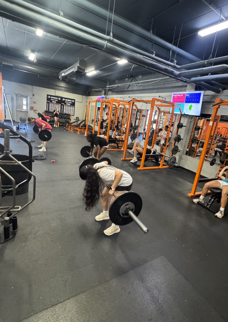 Multiple people in a gym are performing exercises. One woman is bent over, lifting a barbell.