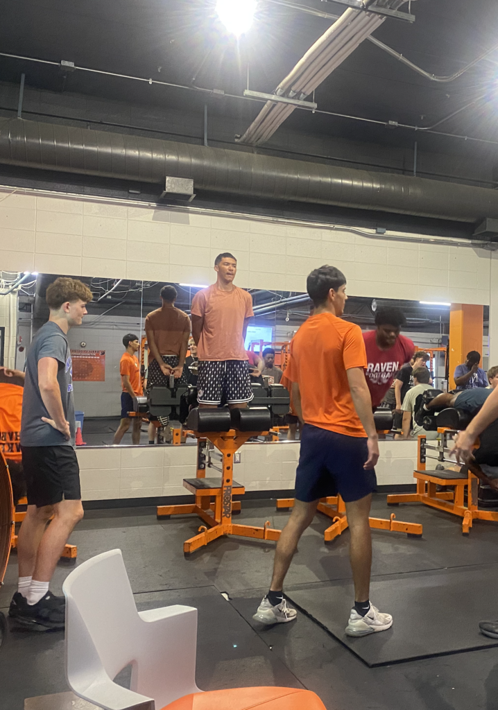 A group of men in various orange tops and shorts in a gym, one on a bench, others standing.