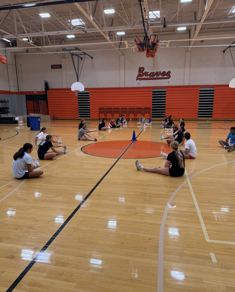 A group of people in a gym, wearing athletic clothing, doing seated exercises in a circle.