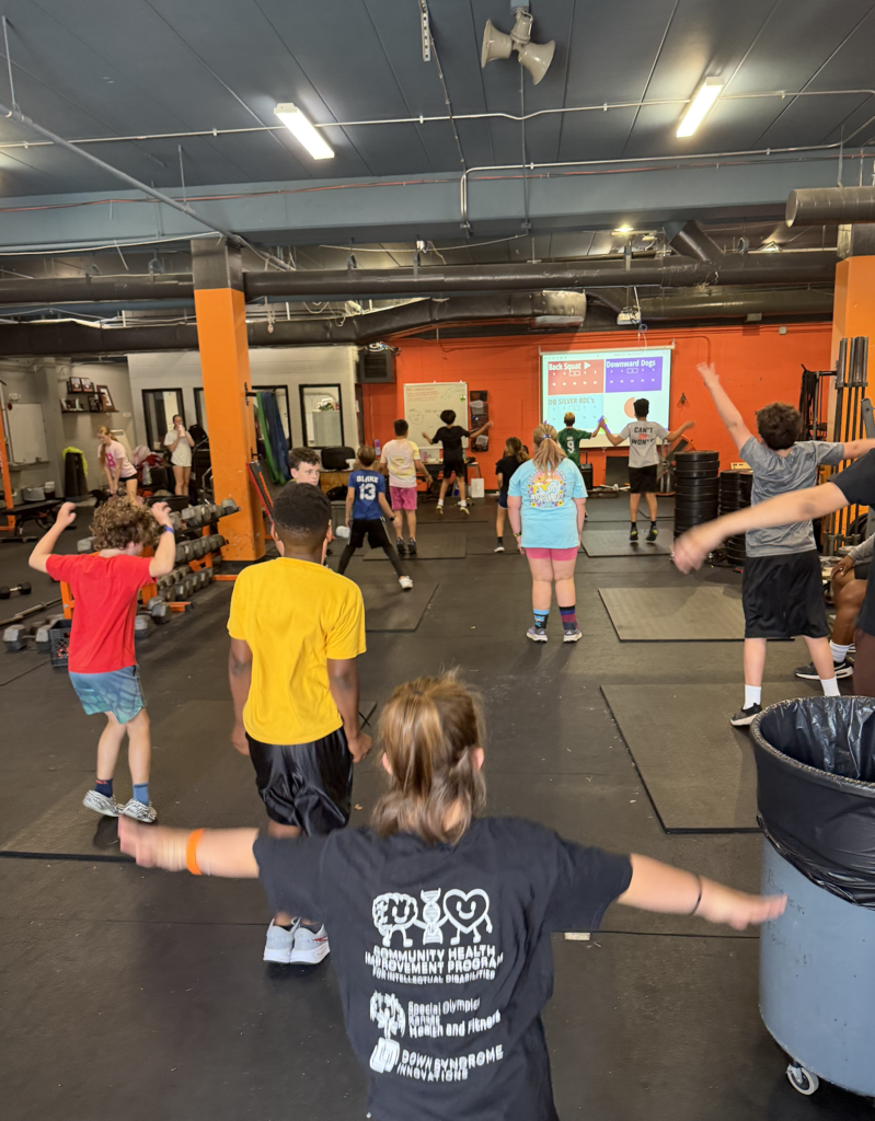 Multiple people in a gym, wearing workout clothes. Some are stretching while others are standing or walking.