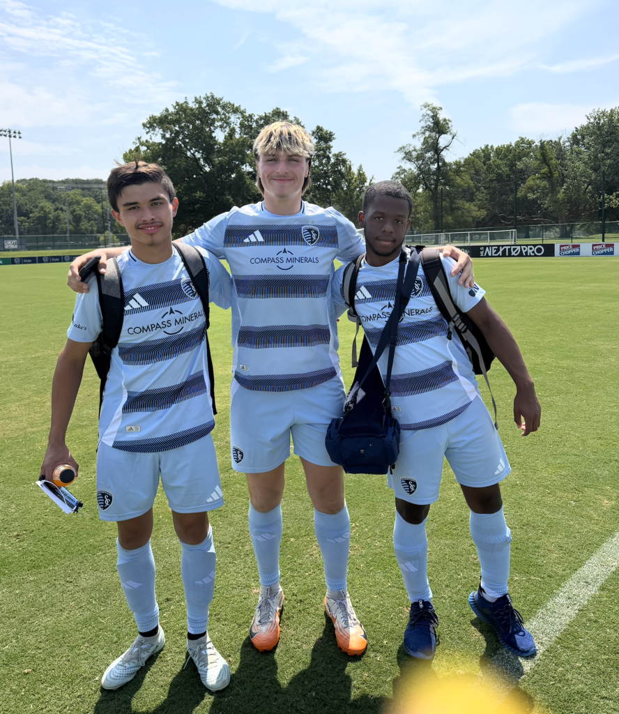 Three soccer players in matching uniforms on a field. Two are standing with their arms around each other.