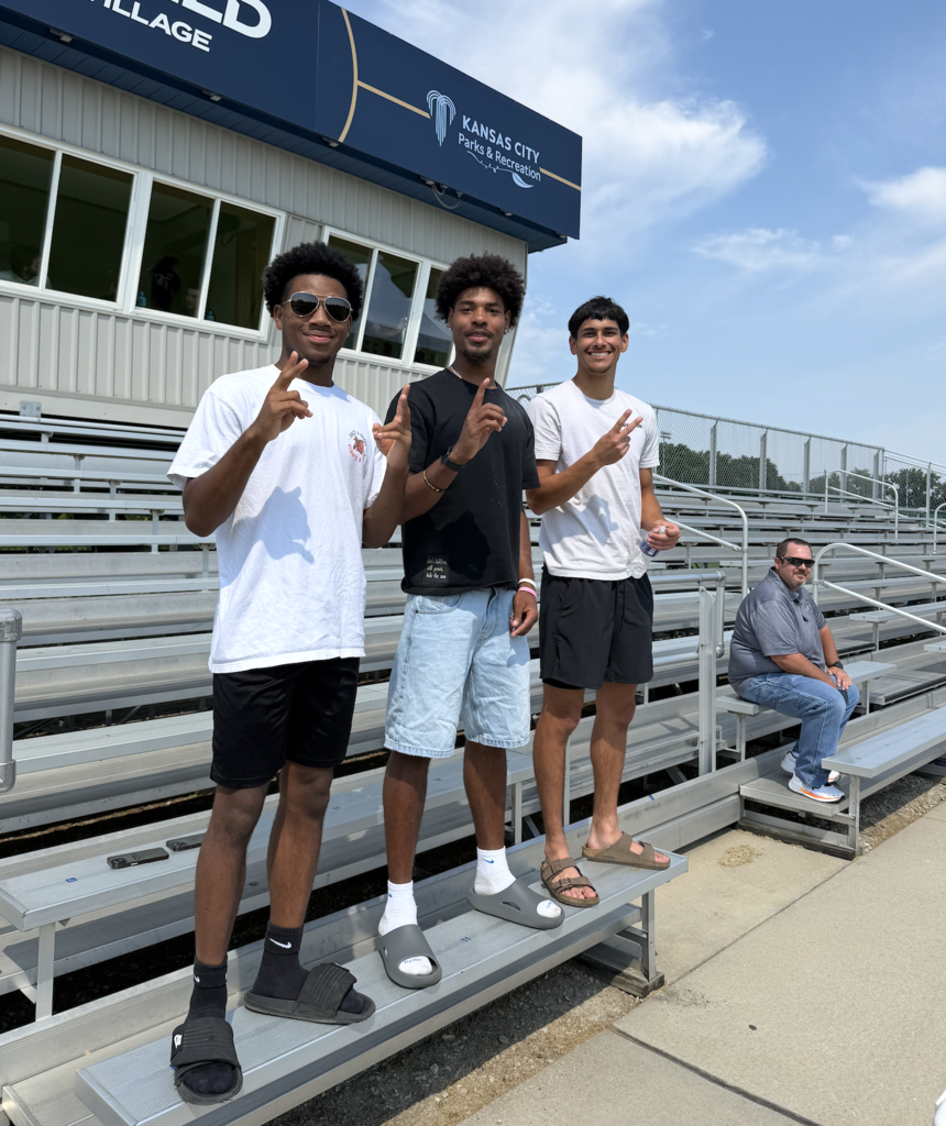 Three boys pose for a photo on bleachers at an outdoor sports facility. A man sits on the lower bleachers.