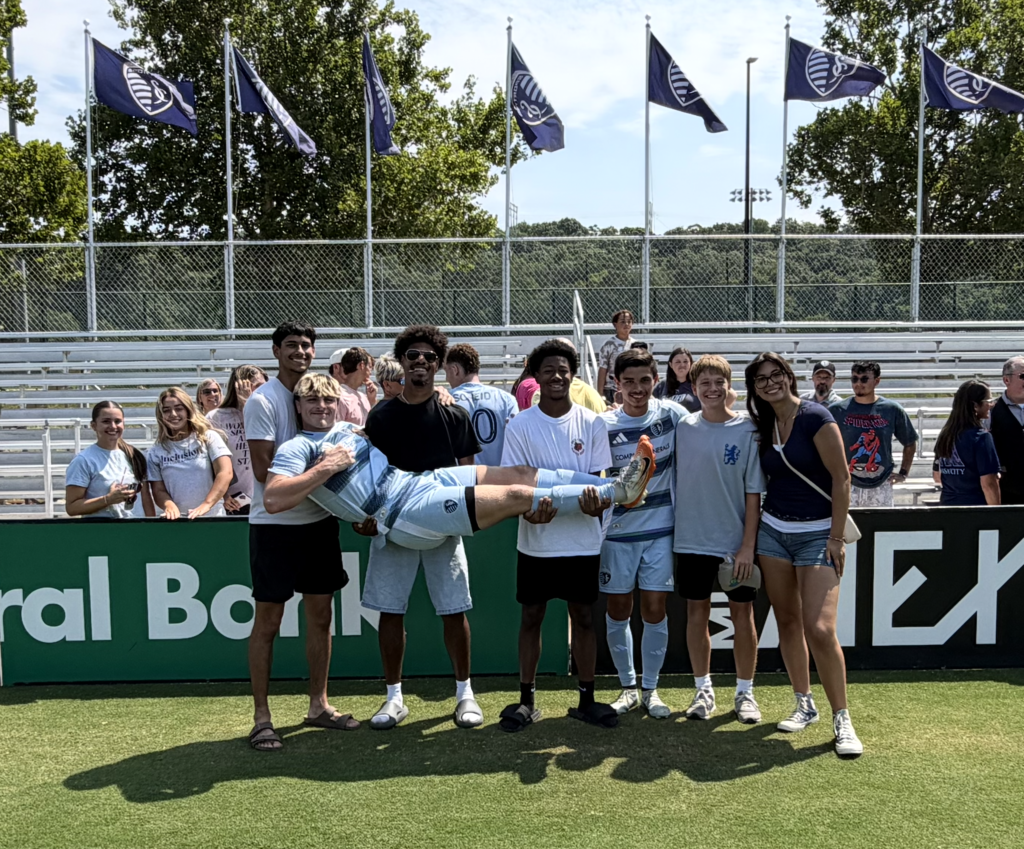 Several people pose for a photo on a sports field with flags and a banner behind them.
