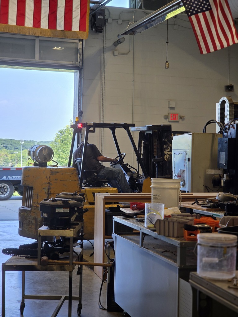 A forklift is positioned inside a workshop. Various equipment and tools are around. Two American flags hang on the walls.