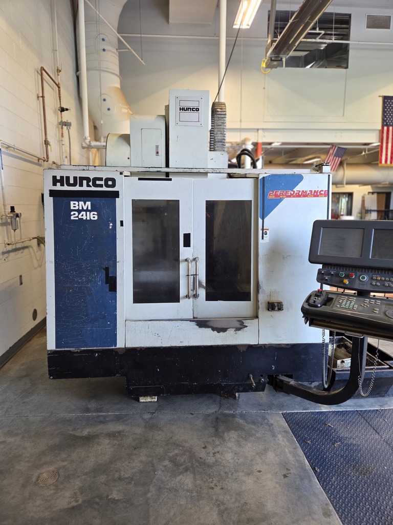 Industrial room with a large white and blue machine labeled "Hurco" on its side, a black floor mat, and an American flag on the wall.