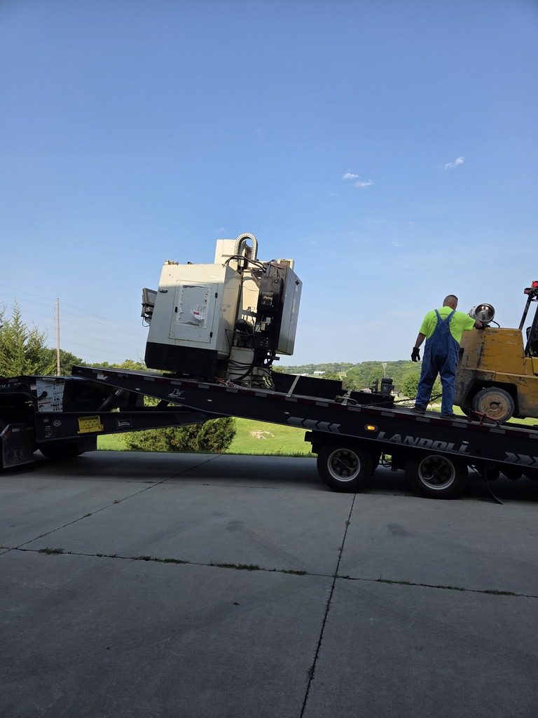 A yellow forklift unloads a large machine from a flatbed trailer in a parking lot.
