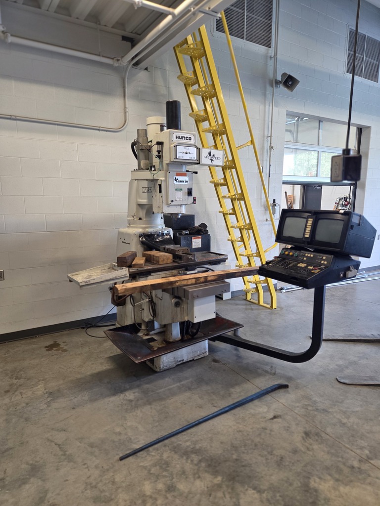 Industrial setting with a large machine and a yellow ladder in the background. The machine has a long table for materials.