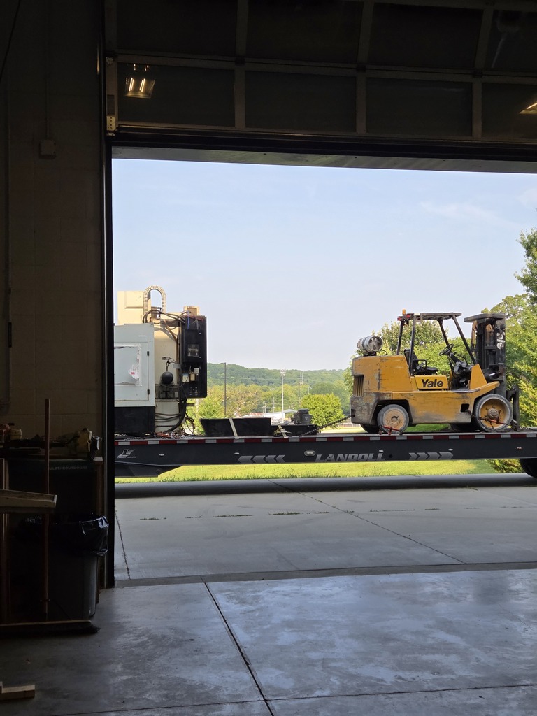 Bright daylight visible through open door of building. Forklift truck loaded with equipment on the road. Trees and sky visible in the distance.