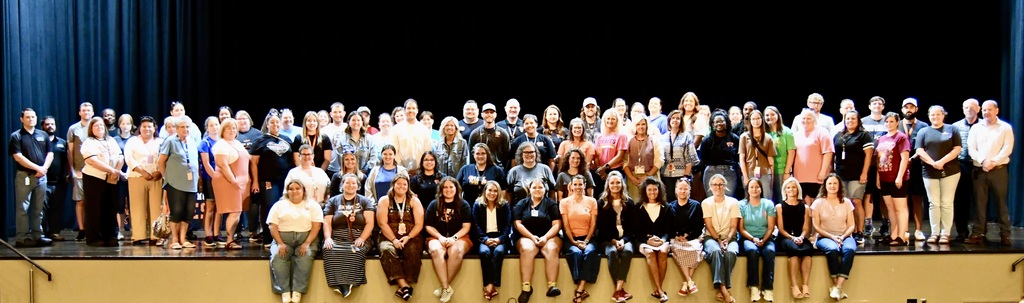A large group of people on a stage, wearing various clothing, and posing for a photo.