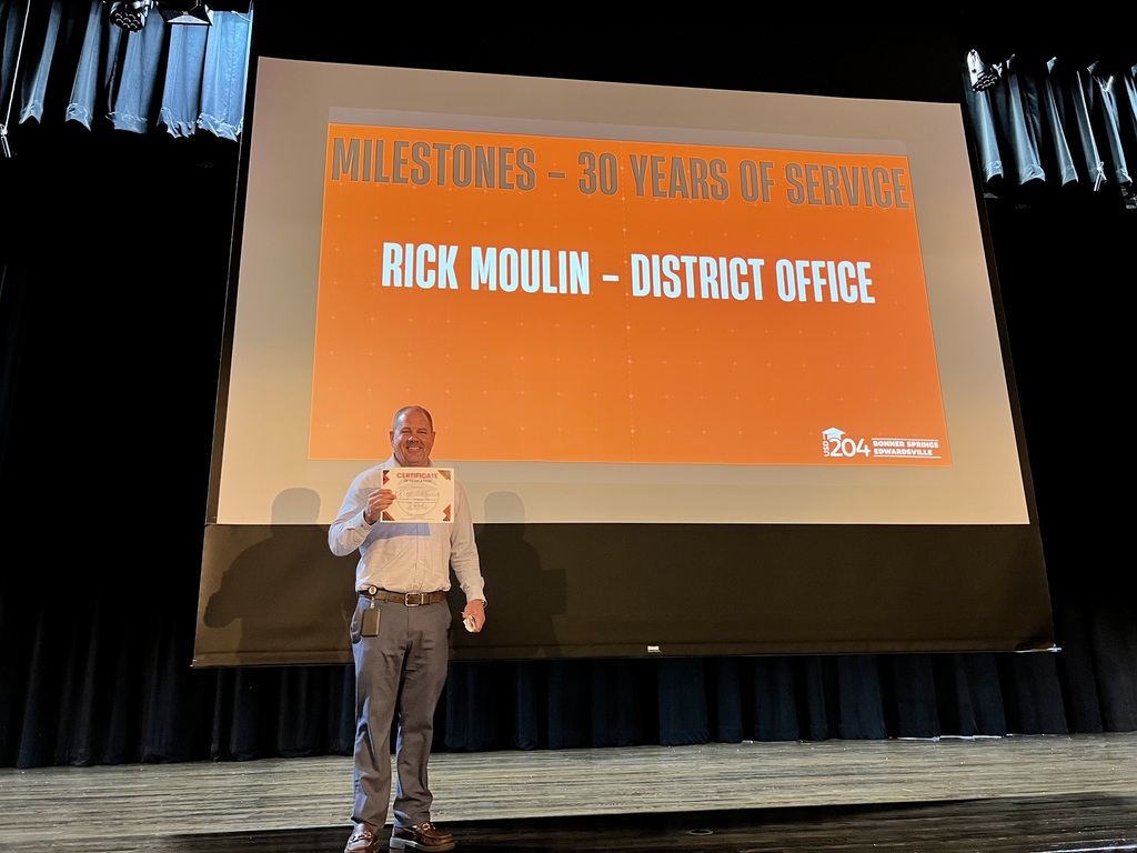 Person standing on stage with a certificate, projector screen behind displaying "Milestones - 30 Years of Service Rick Moulin - District Office."