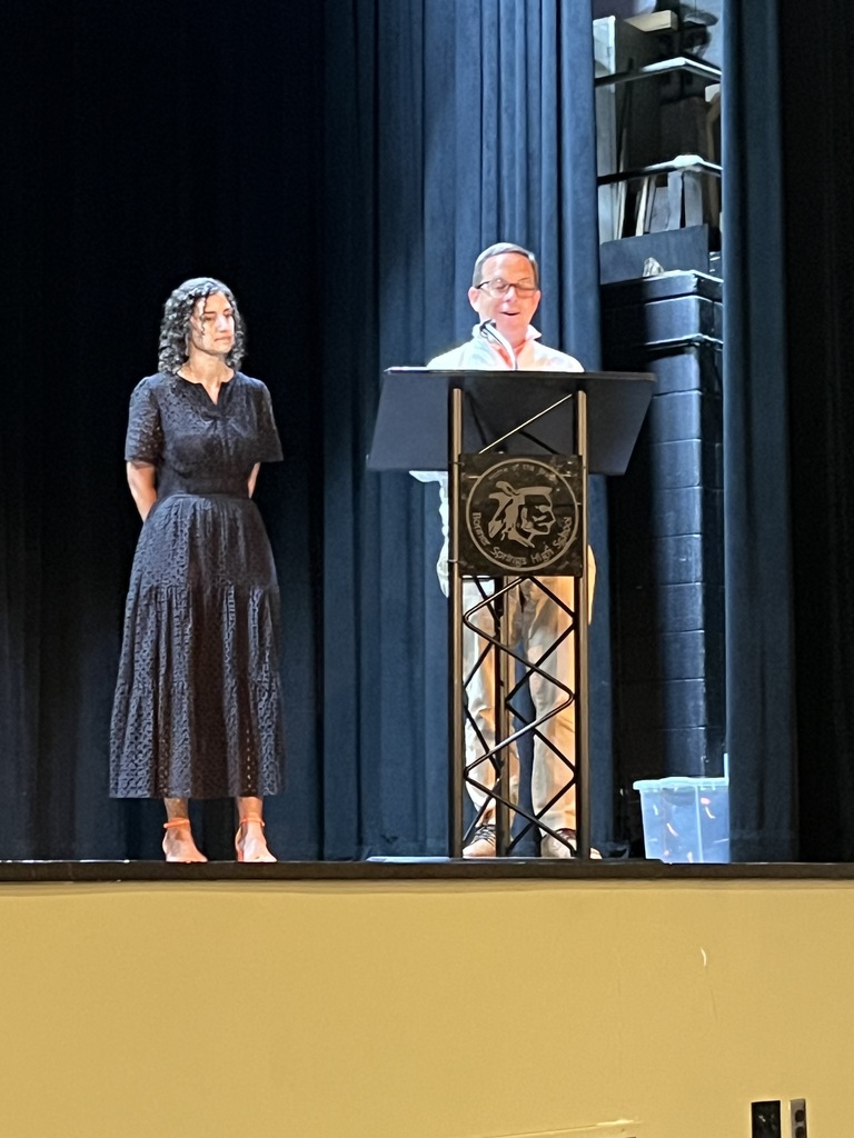 A man and woman on stage with a podium, the man speaking and the woman standing beside.
